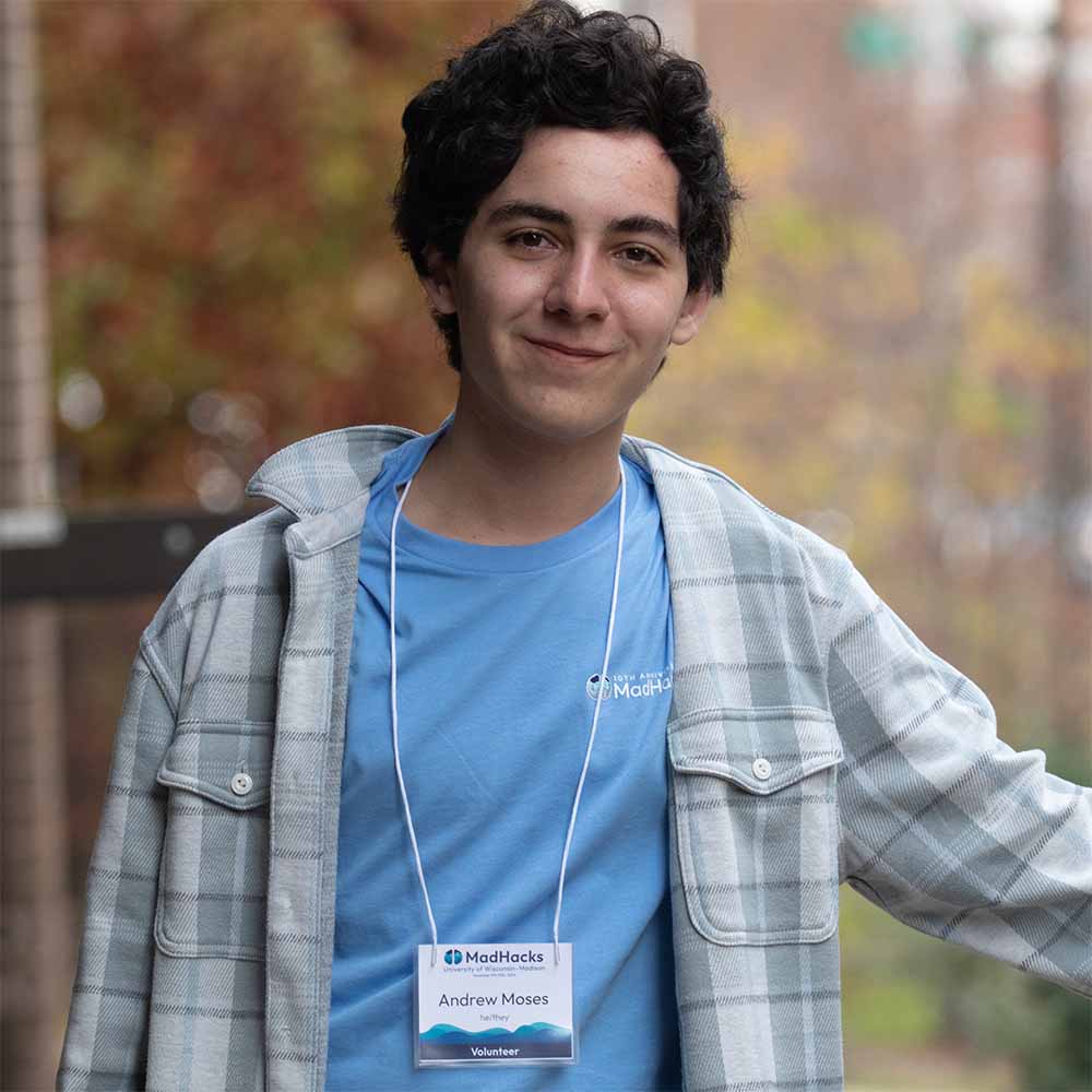 Andrew outside leaning on a fence. He smiles at the camera, wearing a light blue shirt, a gray flannel, and a lanyard with his name on it.