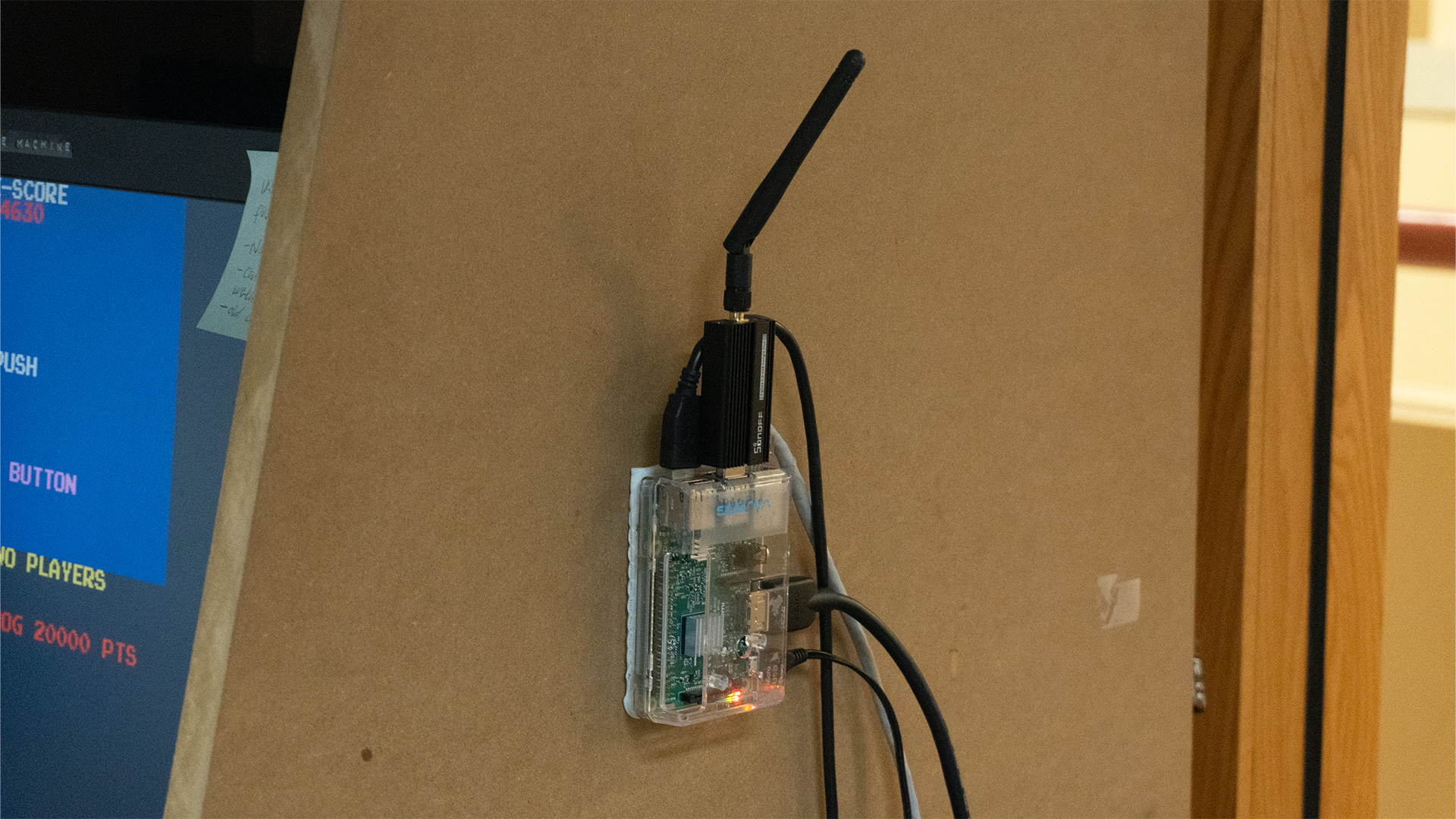 An image of a Raspberry Pi suspended from a wall with various cables plugged into it. There's a USB stick with an antenna sticking out.